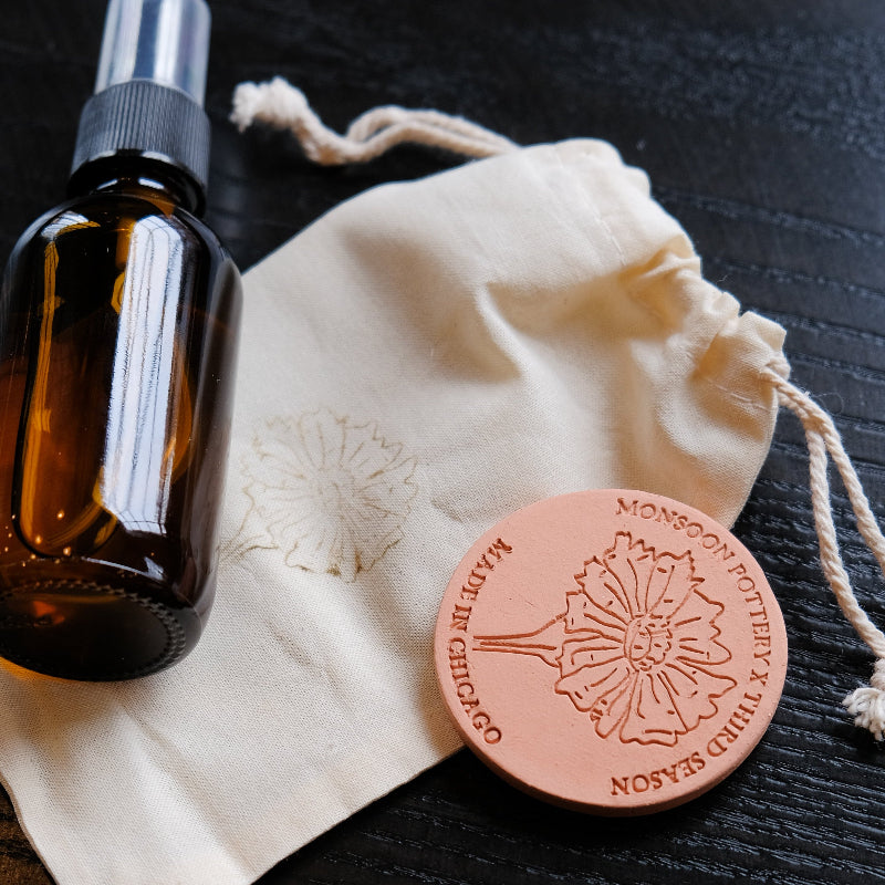 Amber glass bottle with spray nozzle on a white drawstring bag with a terracotta stone diffuser on a dark surface.