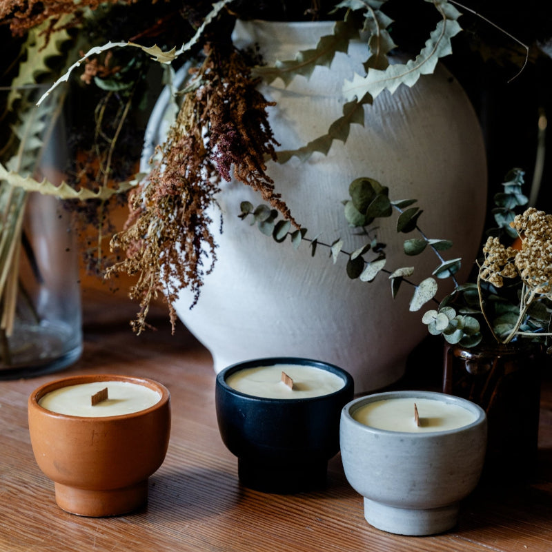 Three candles in different containers on a wooden surface with a vase of dried flowers in the background.